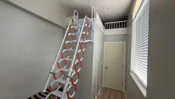 Eine Treppe mit Metallgeländer und orangefarbenen Akzenten, die zu einer Zwischenebene mit einer Tür und einem Fenster mit Jalousien führt.