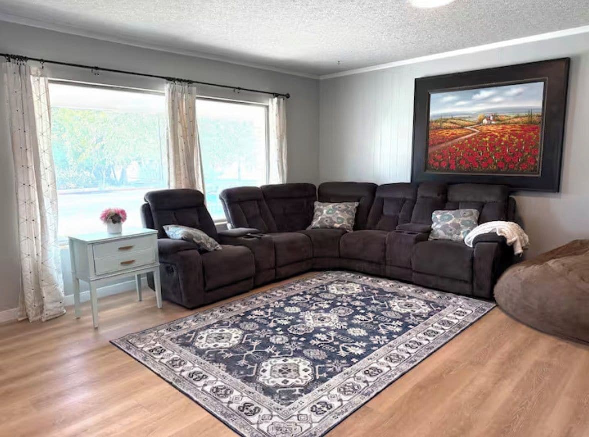A living room with a sectional sofa, a patterned rug, a side table, and a large window with curtains.