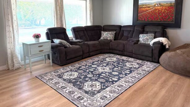 A living room with a sectional sofa, a patterned rug, a side table, and a large window with curtains. - River House (Logan)