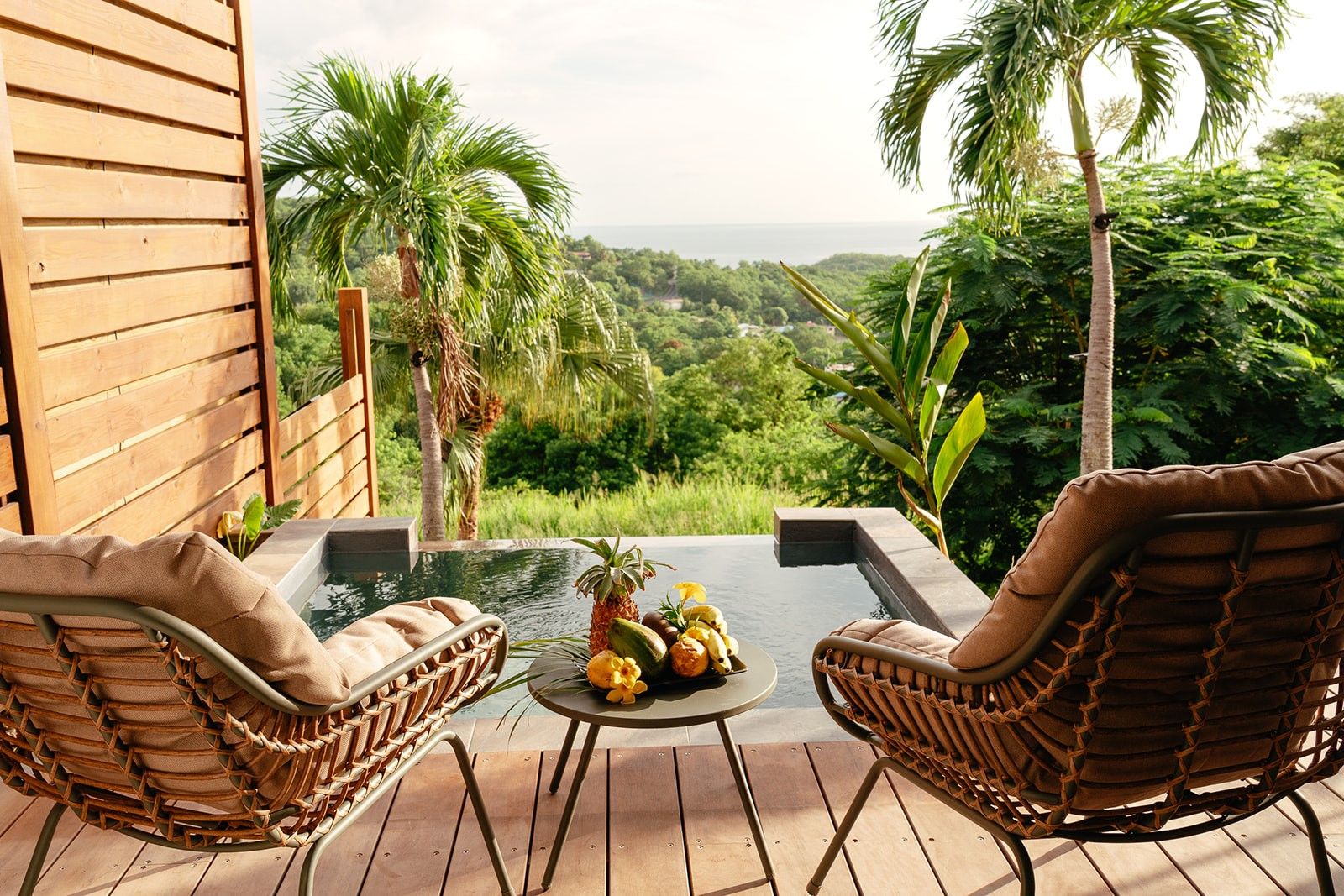 Una terraza de madera con dos sillas de mimbre, una mesita redonda con frutas y vistas a un paisaje exuberante.