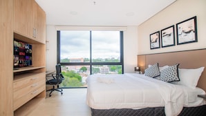 Ein modernes Schlafzimmer mit einem großen Bett, einem Schreibtisch mit Computer, einem Sessel und einem Fenster mit Blick auf die Stadt.