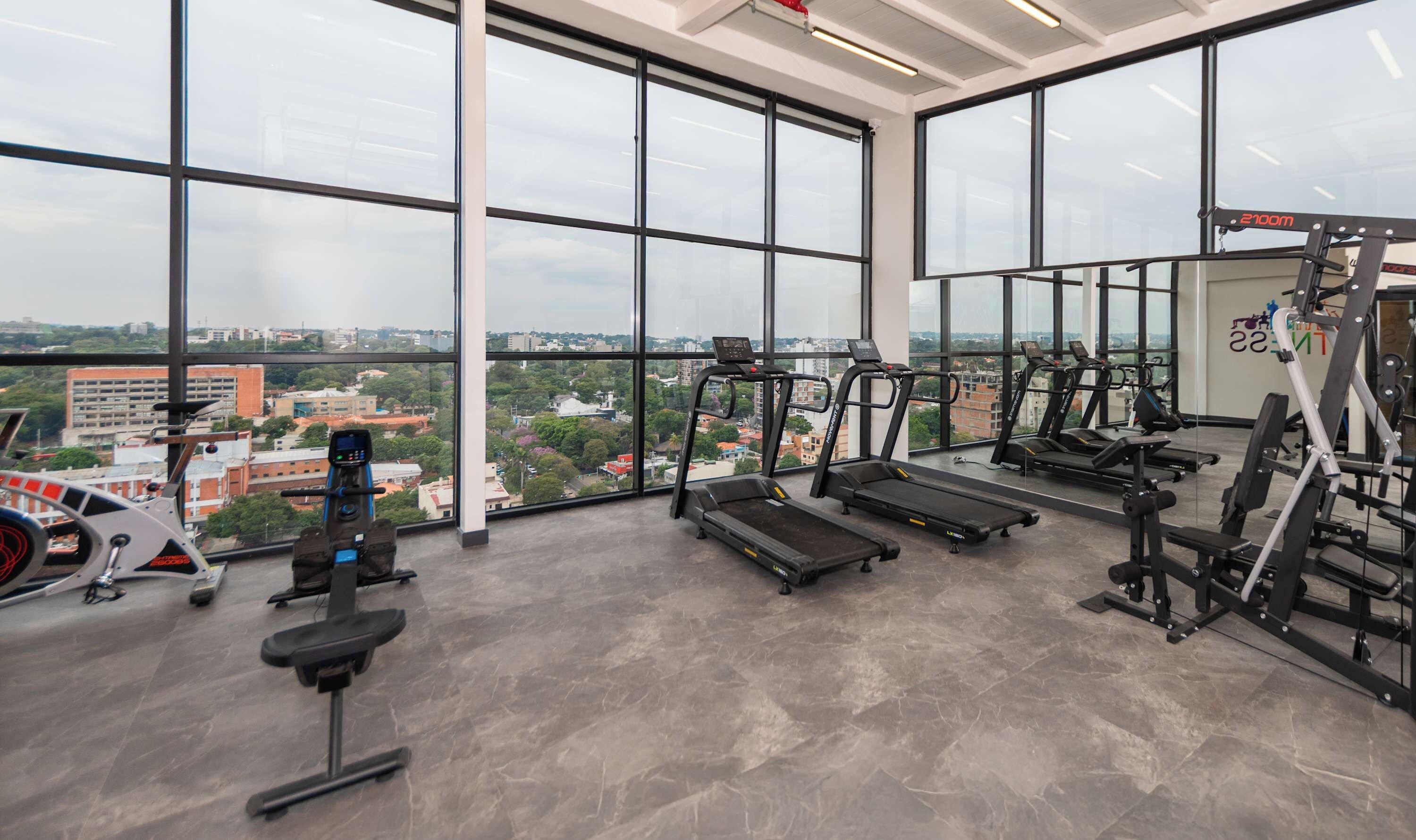 Un gimnasio con cintas de correr, máquinas de pesas y vistas de la ciudad.