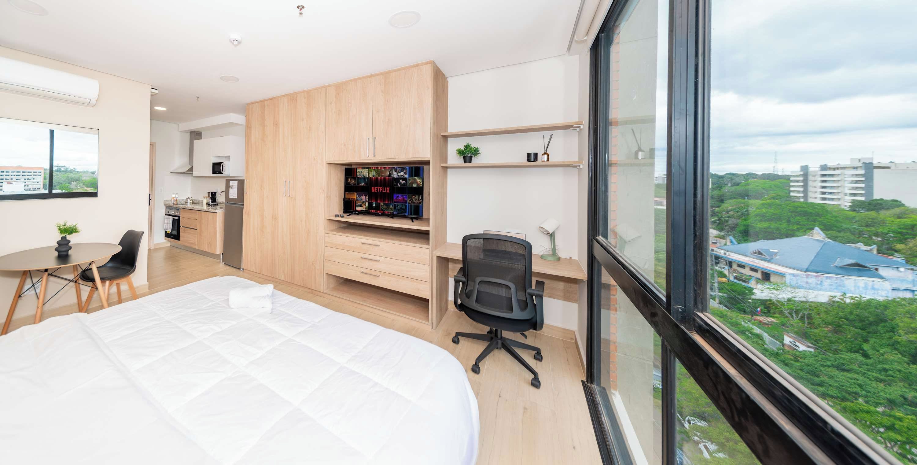 Un dormitorio moderno con una cama grande, un escritorio con silla, kitchenette y un televisor con el logo de Netflix.