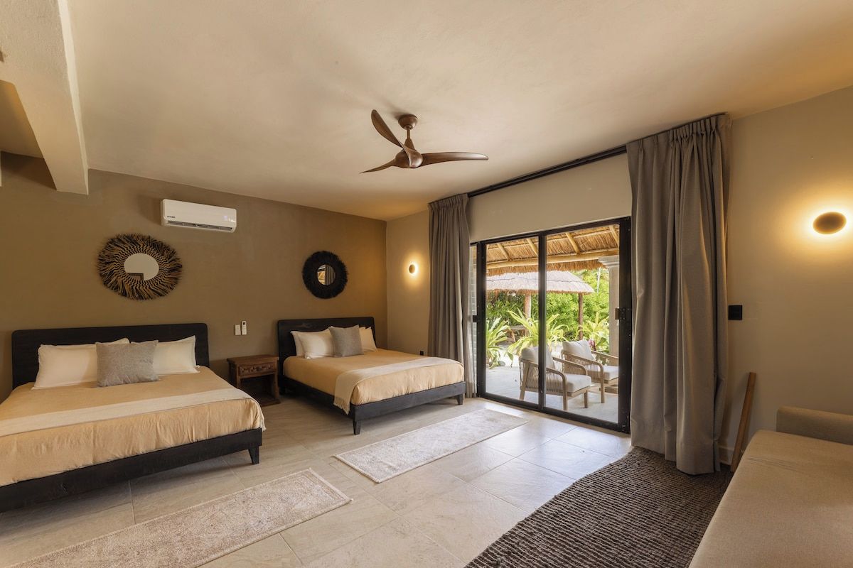 A hotel room with two beds, a ceiling fan, an air conditioning unit, a large sliding glass door to a balcony, and a view of greenery outside.
