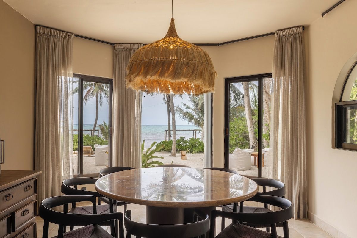 A dining area with a round table and chairs, a hanging light fixture, and a view of the beach through large windows.