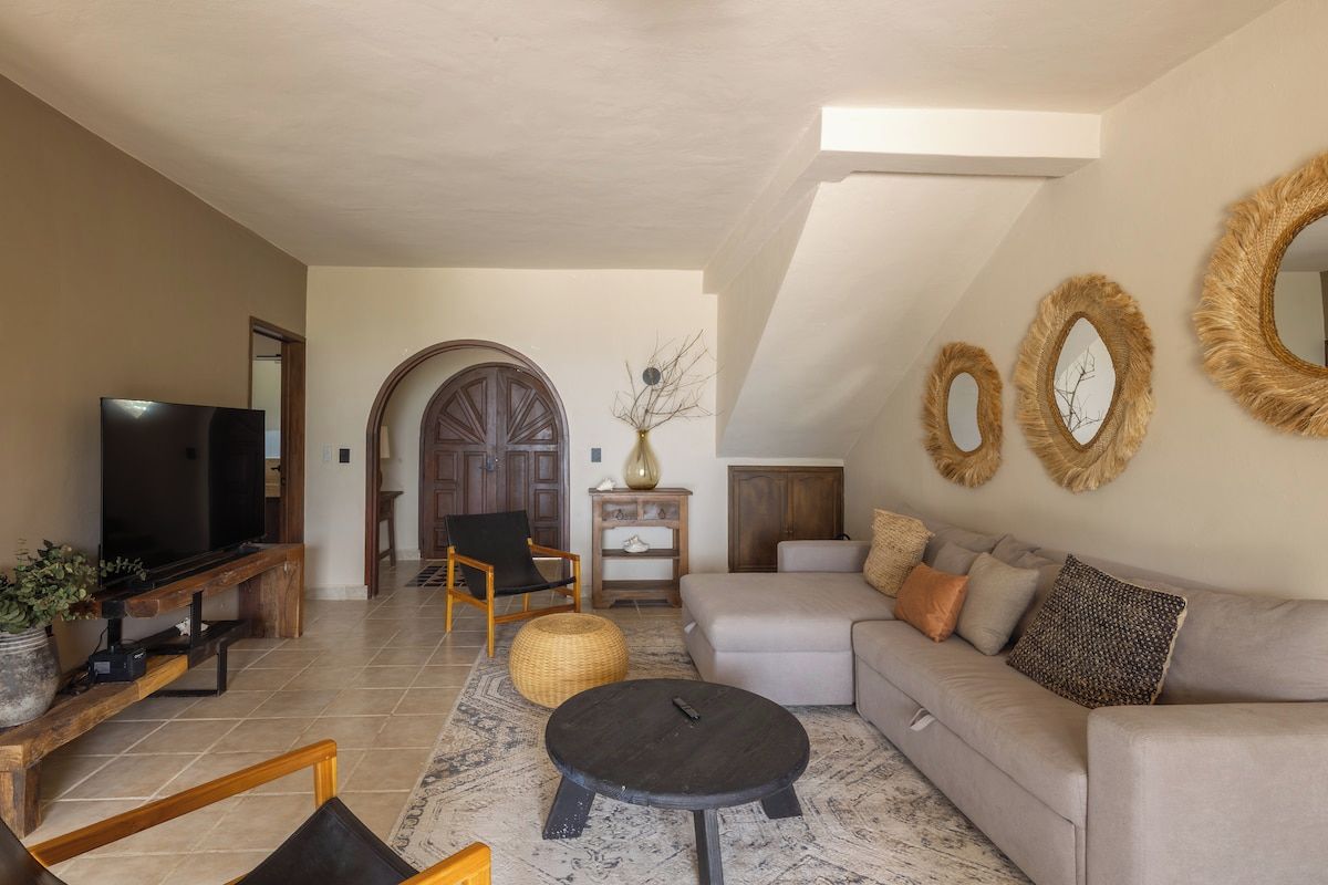 A living room with a television, sofa, and coffee table. There is a wooden bench and a chair. The room has a high ceiling and a sloped roof.