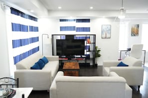 En moderne stue med et fladskærms-TV, hvide sofaer med blå puder, et sofabord og en reol.