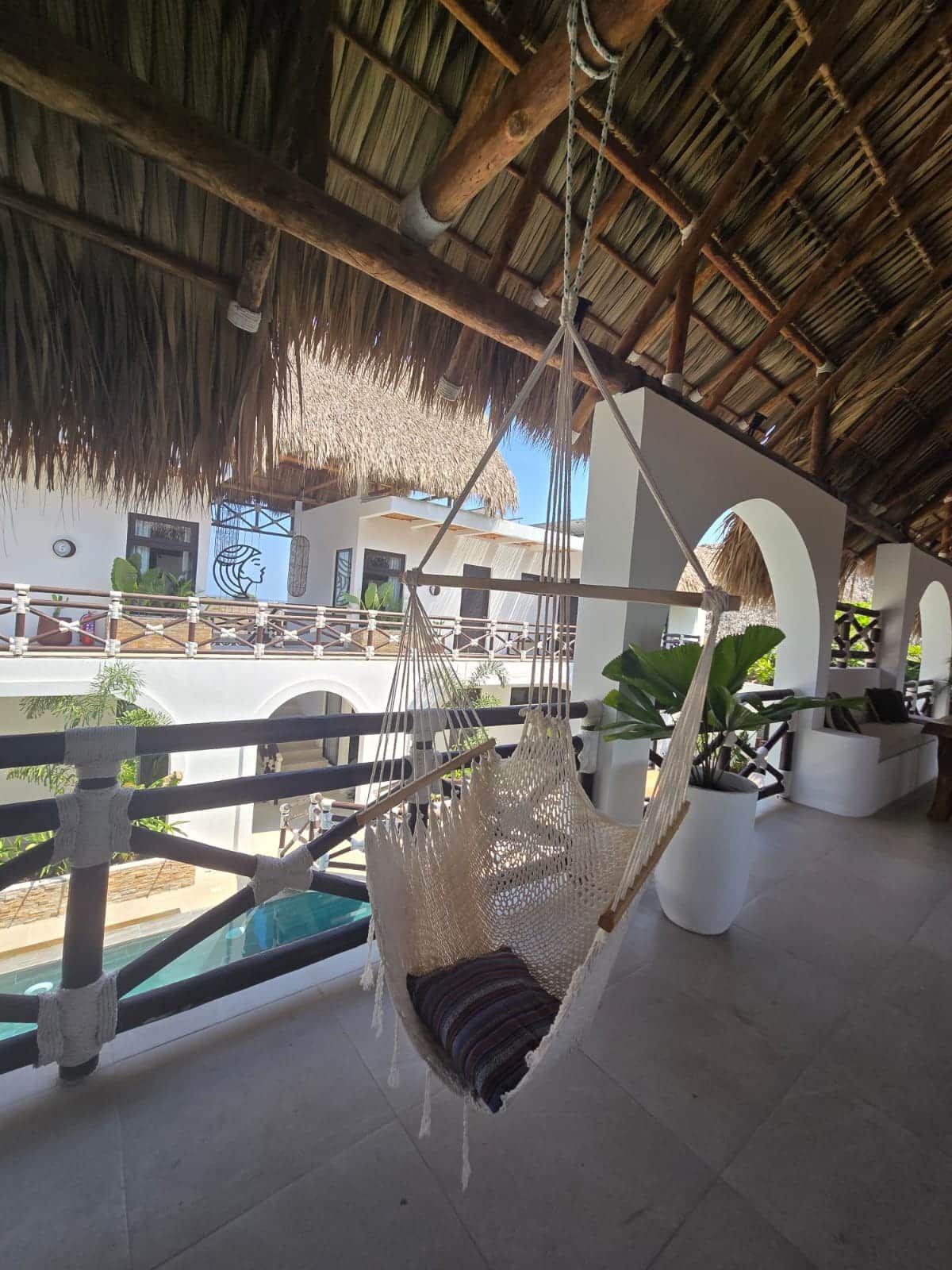 A hammock suspended from a wooden structure, a pool, and a building with arches and a thatched roof.