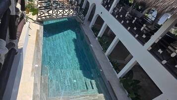 Piscina com deck de madeira e espreguiçadeiras, cercada por paredes brancas e um telhado de palha.
