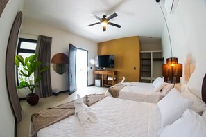 Quarto de hotel com duas camas, ventoinha no teto, televisão, planta e uma janela grande.