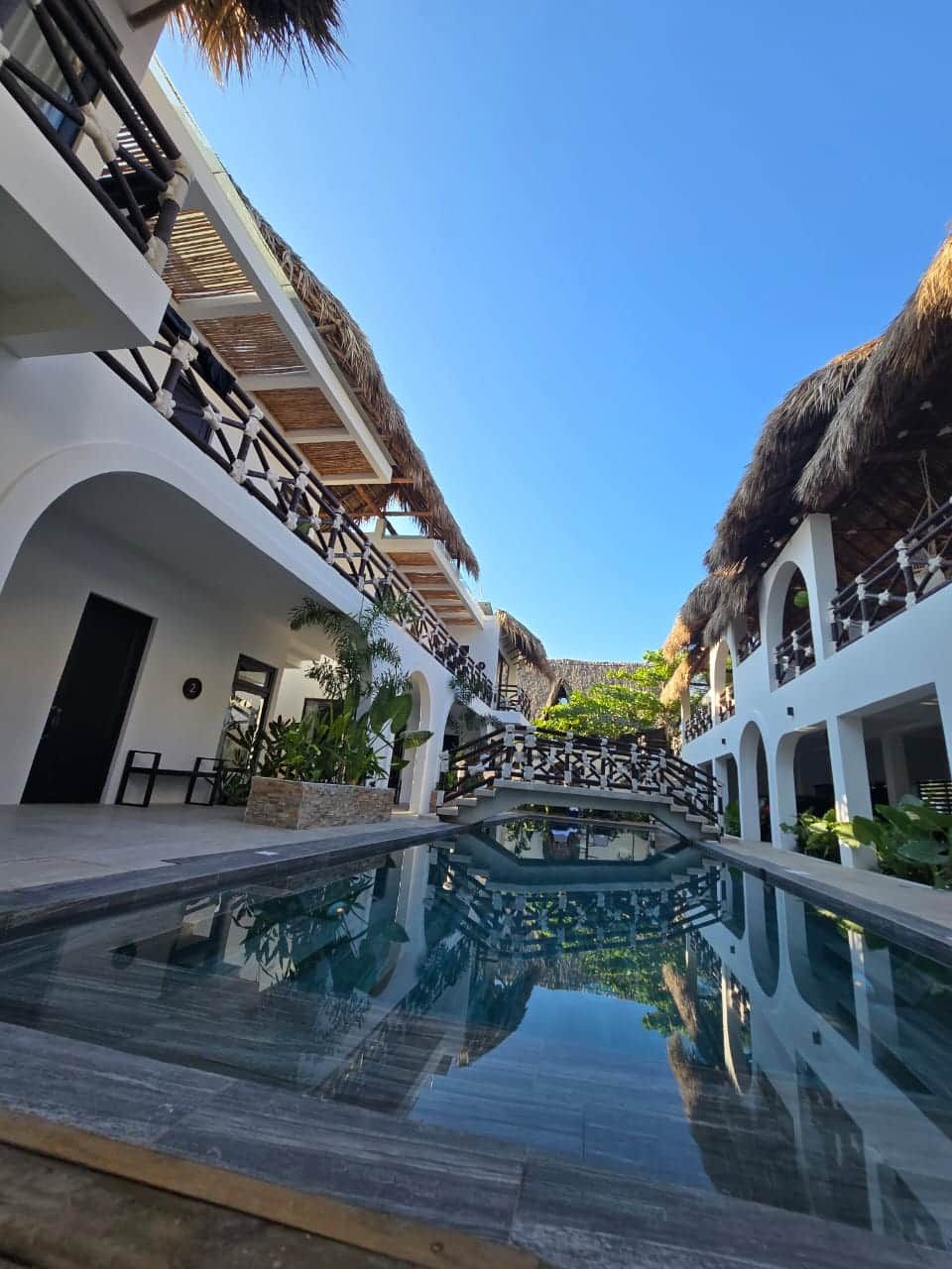 A pool area with a clear view of the sky, surrounded by white buildings with balconies and thatched roofs.