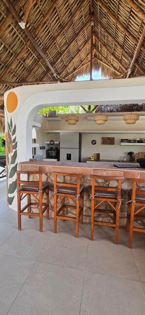 Un área de bar al aire libre con techo de paja, mostrador de piedra y taburetes de madera.