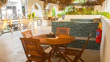 A round wooden table with chairs in a covered outdoor area with a brick wall and a fire extinguisher.