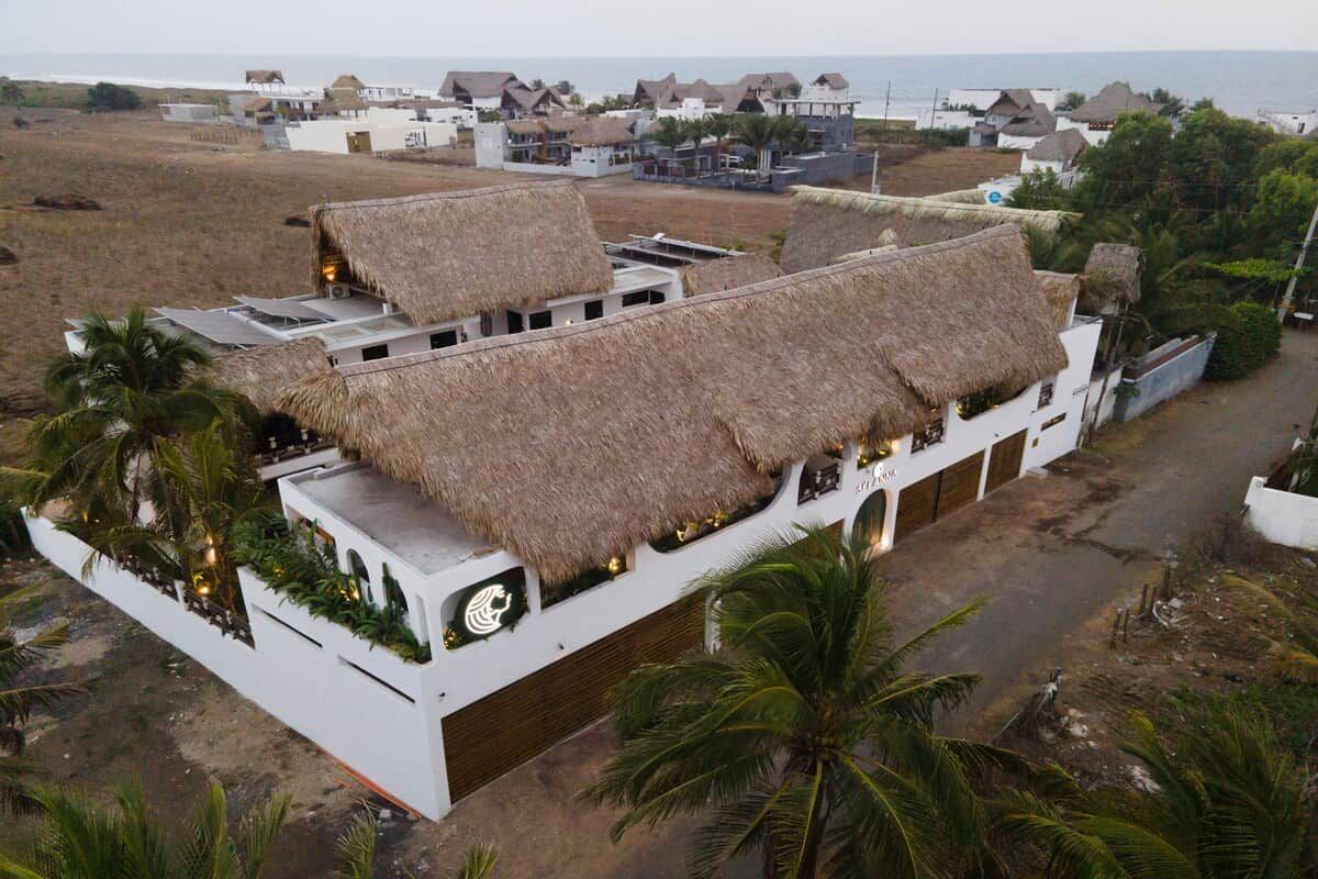 A building with thatched roofs and a white exterior, surrounded by palm trees and a coastal view.