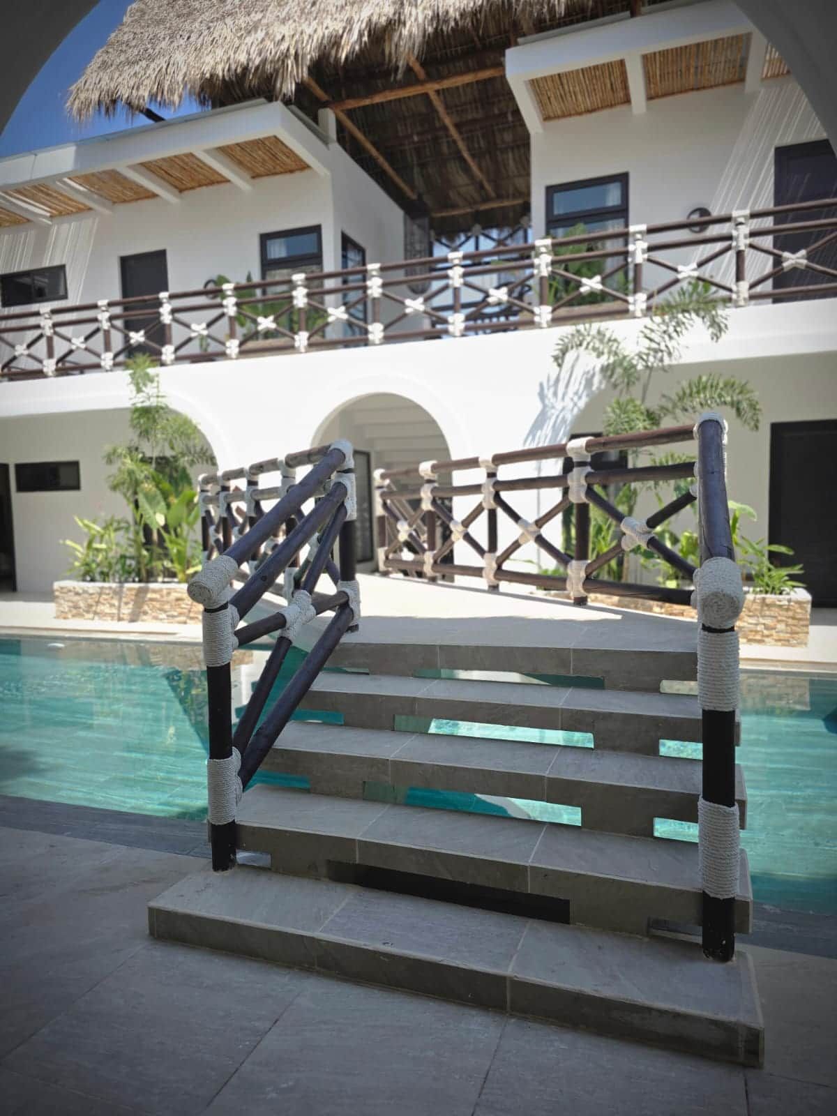 Un área de piscina con una escalera que lleva a un balcón, rodeada por un edificio blanco de techo de paja.