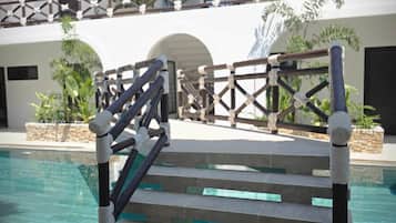 Área de piscina com escadaria para uma varanda, cercada por um edifício branco com telhado de palha.