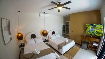 Quarto de hotel com duas camas, um ventilador de teto, uma televisão e uma pequena mesa de cabeceira de madeira.