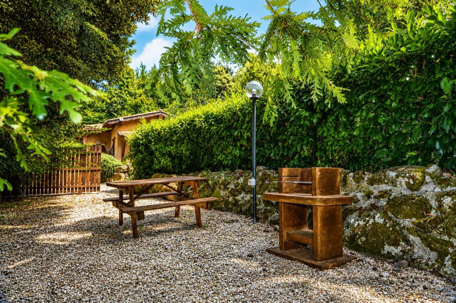 Une terrasse en gravier avec une table et des bancs en bois, entourée d’un feuillage dense et d’un mur de pierre.