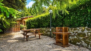 Um pátio de cascalho com mesa e bancos de madeira, rodeado por folhagem densa e uma parede de pedra.