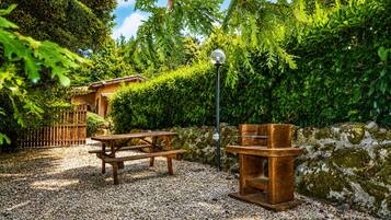 Une terrasse en gravier avec une table et des bancs en bois, entourée d’un feuillage dense et d’un mur de pierre.