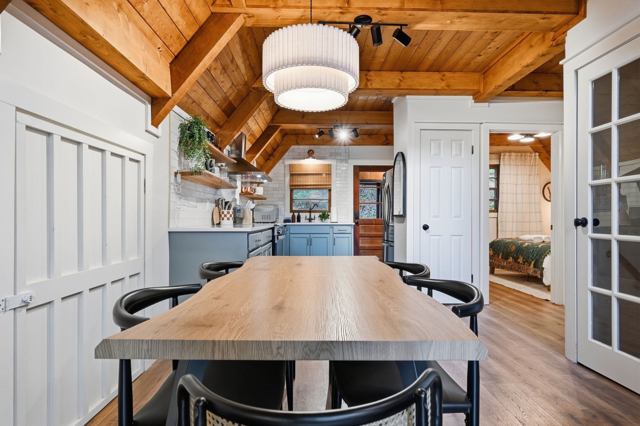 A modern kitchen with a wooden table and chairs, a hanging light fixture, and a view into a bedroom.