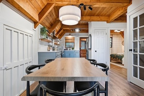 A modern kitchen with a wooden table and chairs, a hanging light fixture, and a view into a bedroom. - Dutch Creek A-Frame | Hot Tub, Fire Pit, Trailhead (Banner Elk)