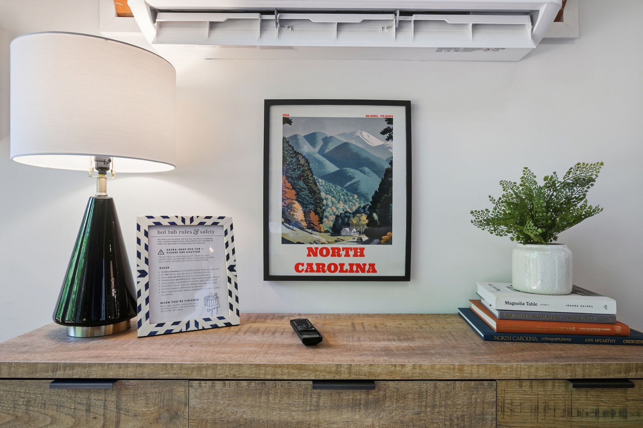 A framed poster of North Carolina, a lamp, a framed set of hot tub rules, a remote control, and a plant on a wooden surface.