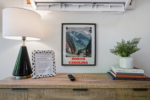A framed poster of North Carolina, a lamp, a framed set of hot tub rules, a remote control, and a plant on a wooden surface. - Dutch Creek A-Frame | Hot Tub, Fire Pit, Trailhead (Banner Elk)