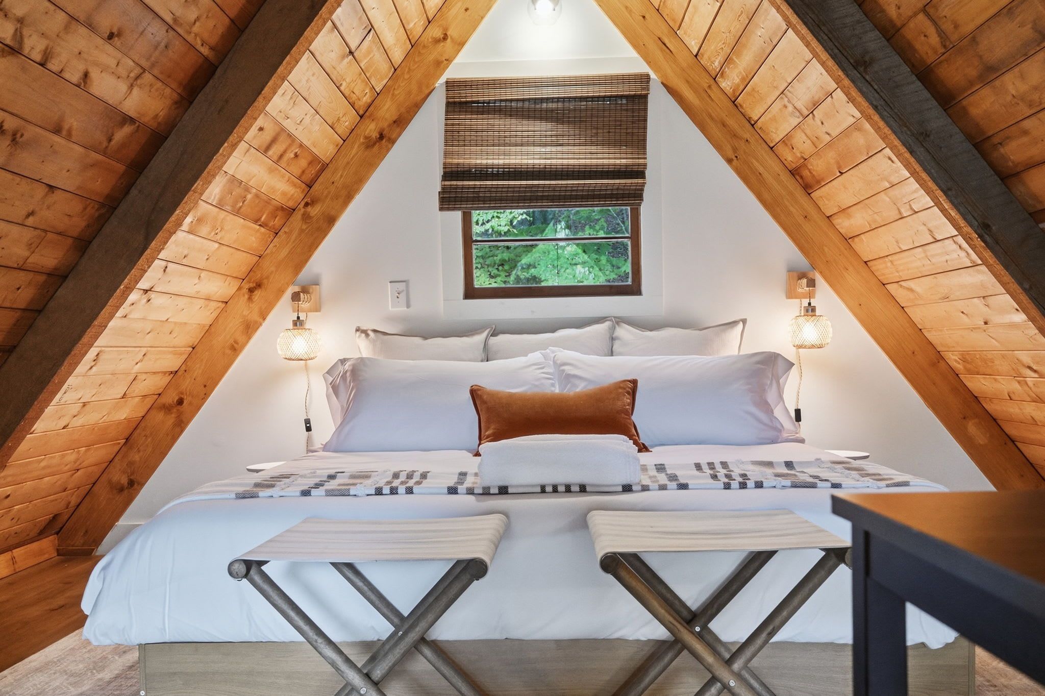 A bedroom with a sloped ceiling, a bed with white bedding, two bedside lamps, and a window with a view of greenery.