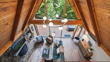 A high-ceilinged wooden interior with a stone fireplace, a sofa, and a dining area with outdoor views.