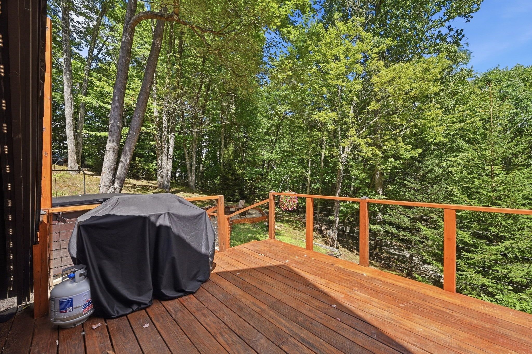 A wooden deck with a barbecue grill and a propane tank, surrounded by a wooden railing and dense forest.