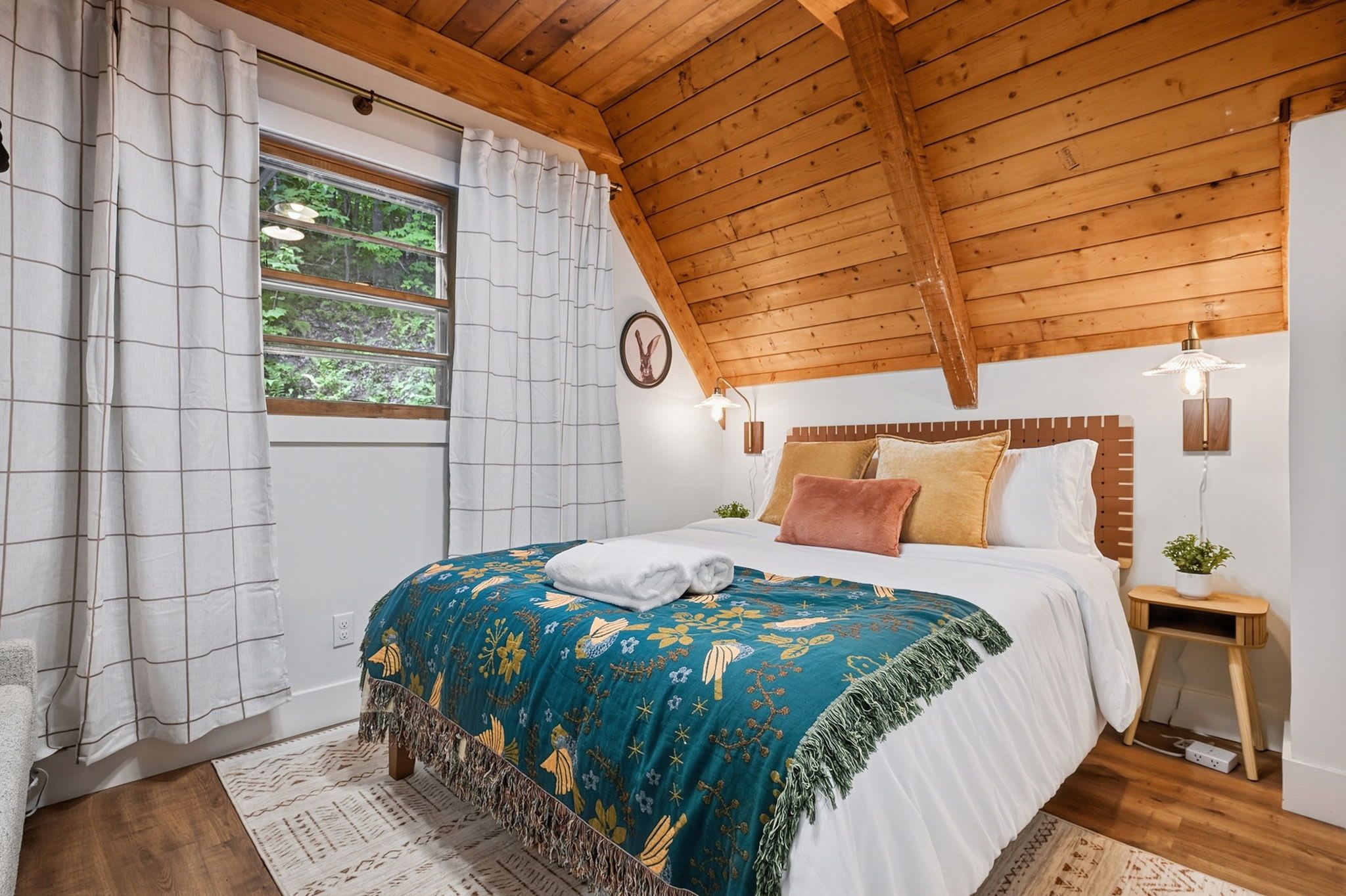 A bedroom with a wooden ceiling, a bed with a blue floral bedspread, a white pillow, and a nightstand with a lamp.