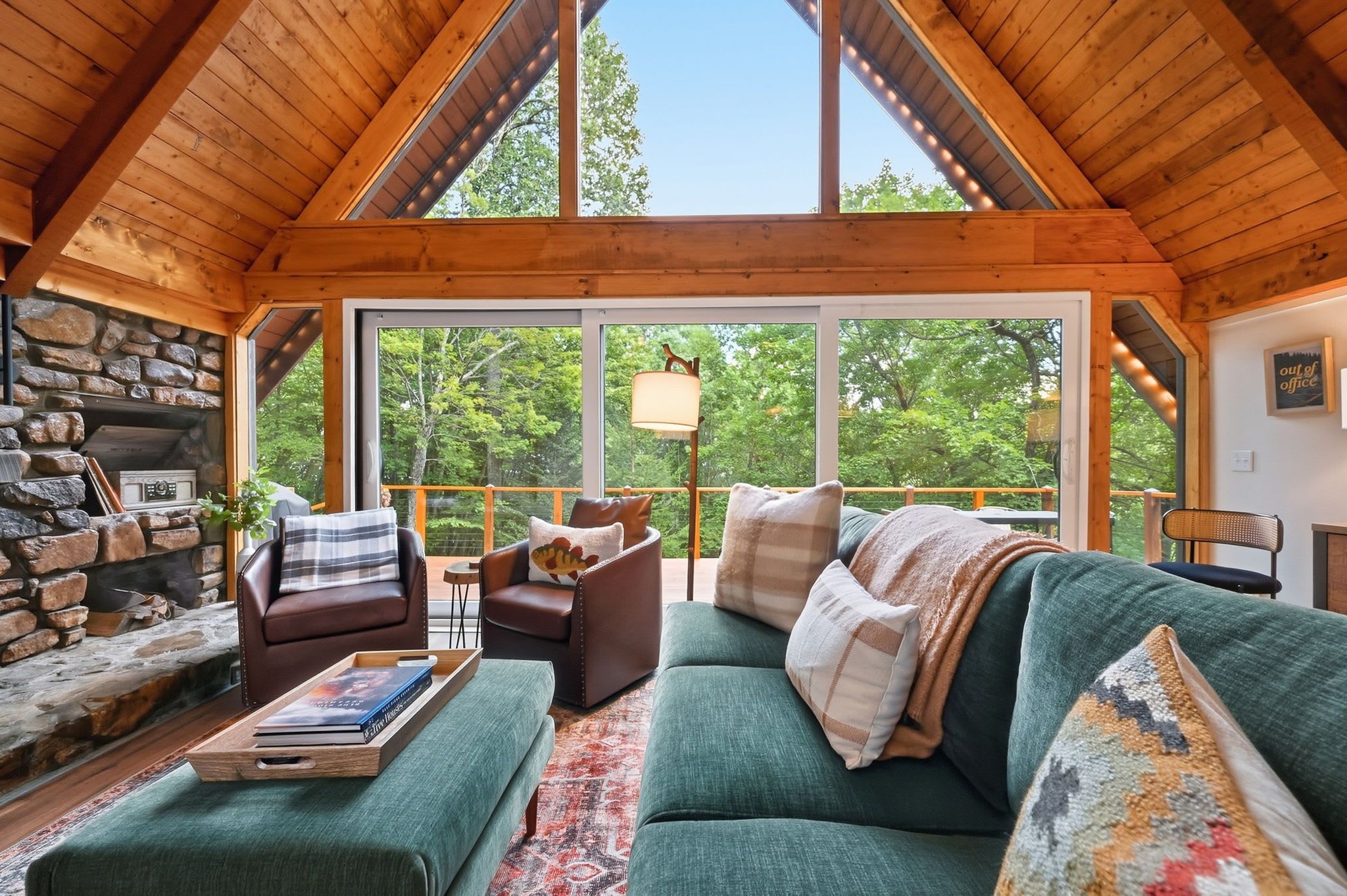 A cozy cabin interior with a stone fireplace, a green sofa, and a large window offering a view of the forest.
