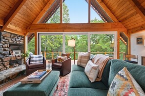 A cozy cabin interior with a stone fireplace, a green sofa, and a large window offering a view of the forest. - Dutch Creek A-Frame | Hot Tub, Fire Pit, Trailhead (Banner Elk)
