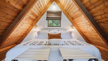 A bedroom with a sloped ceiling, a bed with white bedding, a window with a view of greenery, and a wall-mounted light fixture.