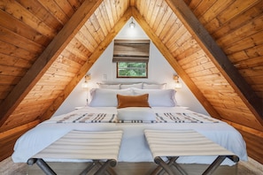 A bedroom with a sloped ceiling, a bed with white bedding, a window with a view of greenery, and a wall-mounted light fixture.