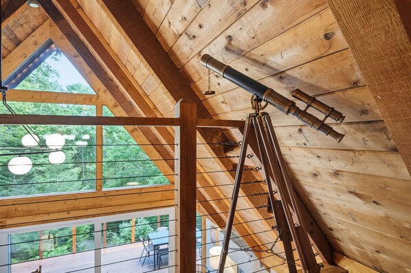 A wooden interior with a telescope mounted on a tripod, large windows, and a view of greenery outside.