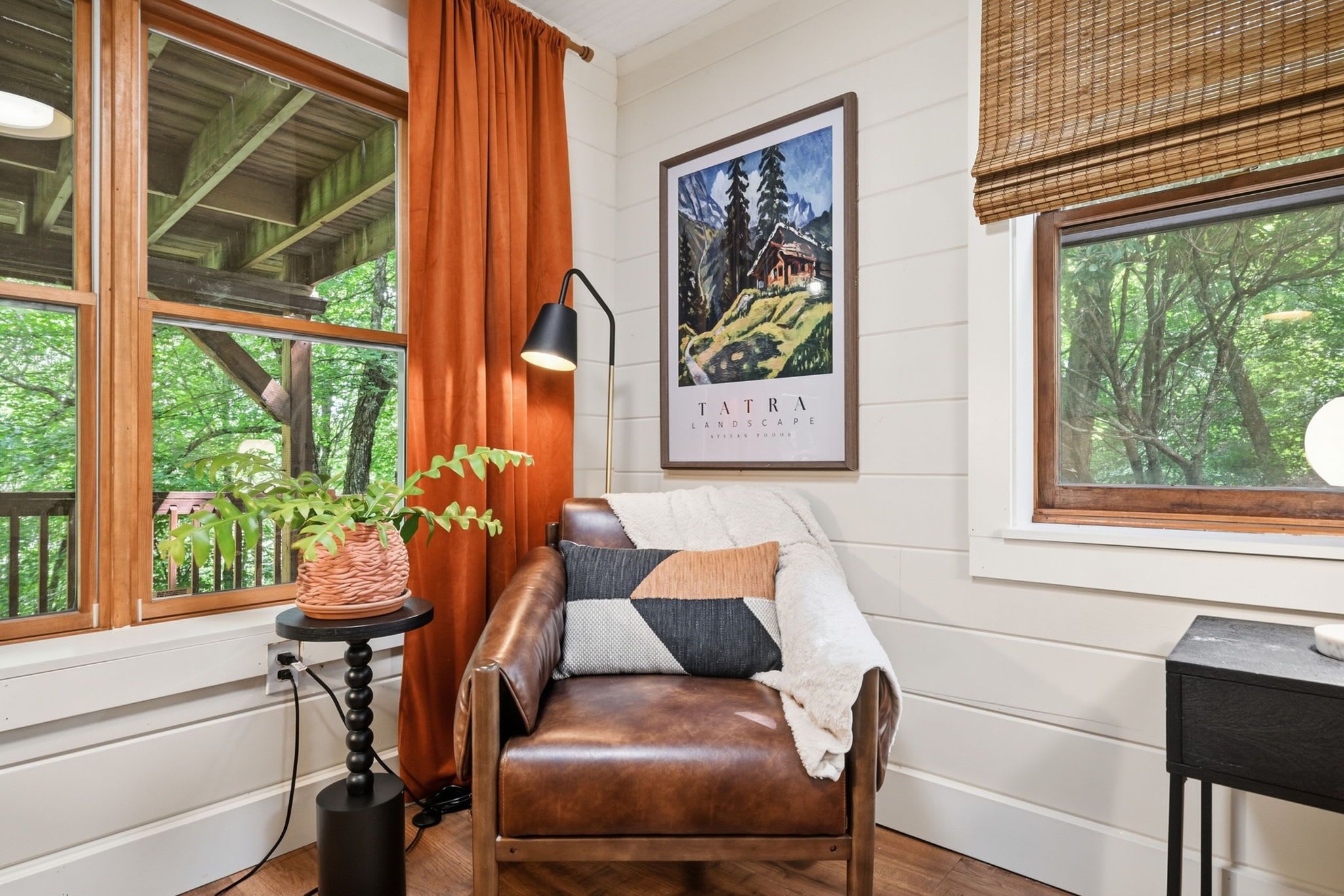 A cozy room with a leather sofa, a framed landscape painting, and a view of trees through the window.