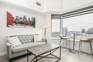A modern living room with a grey sofa, a coffee table, and a city view through large windows. - Modern 1BR with Breathtaking Yorkville View (Toronto)
