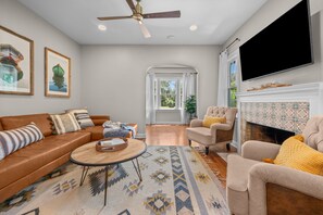A living room with a brown leather sofa, a beige armchair, a round coffee table, and a television mounted on the wall. - Historic 4BR Cottage | Near Downtown and Beach (St. Augustine)