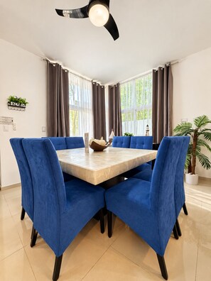 A dining area with a round table, blue upholstered chairs, a ceiling fan, and large windows with curtains. - Luxury family villa with a private pool, spacious rooms, and top-notch amenities (Puerto Morelos)