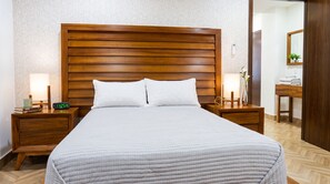 Ein Hotelzimmer mit einem großen Bett, einem hölzernen Kopfteil, zwei Nachttischen mit Lampen, einem Beistelltisch, einem Spiegel und einem kleinen Schreibtisch mit Lampe.