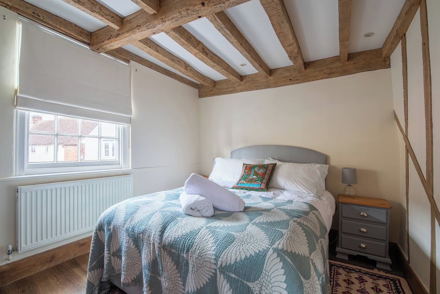 A bedroom with a bed, a nightstand, a window, and exposed wooden beams.