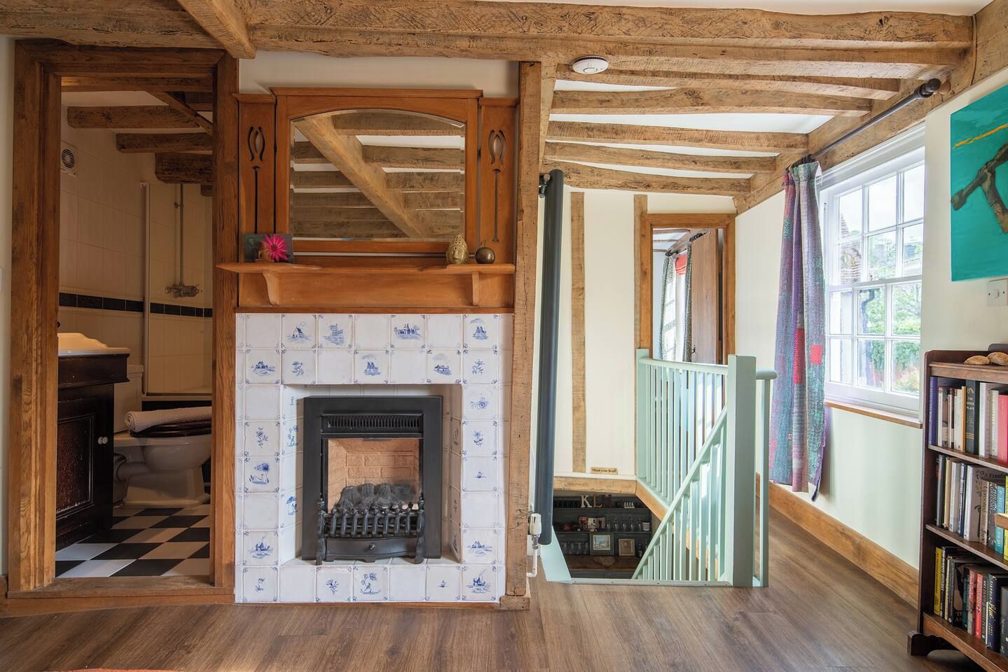 A room with a fireplace, wooden beams, a staircase, and a bookshelf.