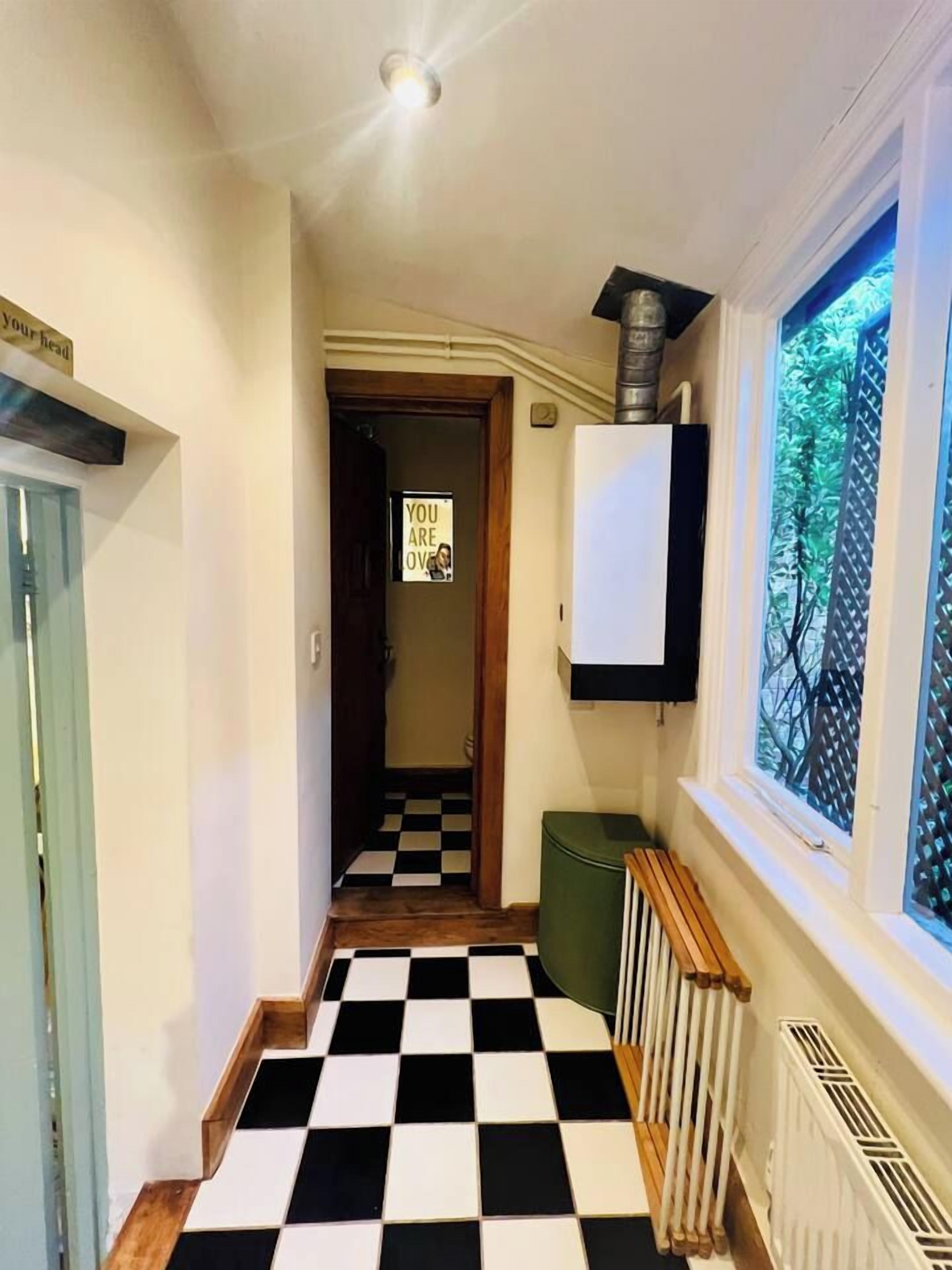 Un pasillo con piso a cuadros, un radiador, una ventana con vista a árboles y una puerta con un letrero.