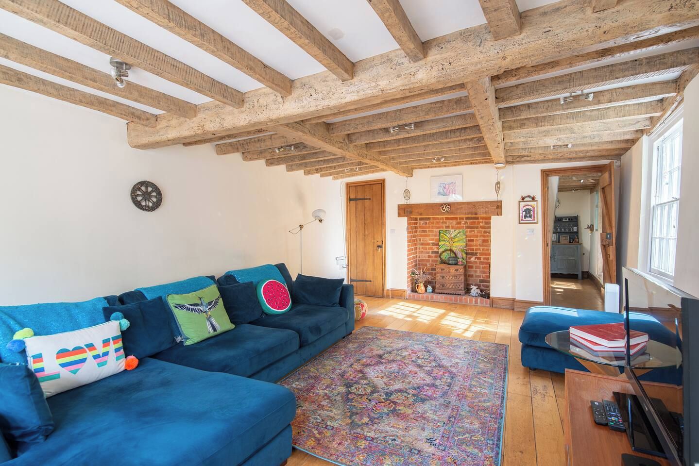 A room with a blue sofa, a patterned rug, wooden beams on the ceiling, a fireplace, and a television on a wooden table.