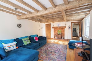 Ein Zimmer mit einem blauen Sofa, einem gemusterten Teppich, Holzbalken an der Decke, einem Kamin und einem Fernseher auf einem Holztisch.
