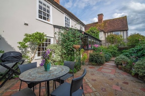 A patio area with a round table and chairs, surrounded by lush greenery and a brick pathway.