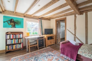Ein Zimmer mit freiliegenden Holzbalken, einem Schreibtisch mit Stuhl, einem Bücherregal, einem Fernseher und einem Bett mit gemustertem Überwurf.