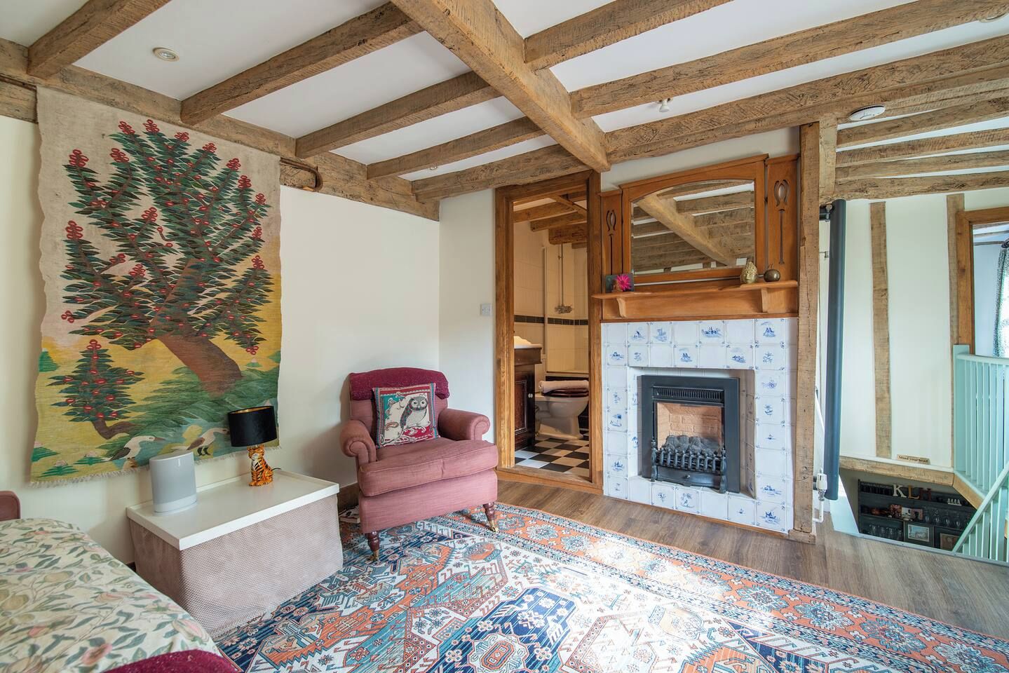 Une pièce avec des poutres apparentes, une cheminée, un tapis à motifs et un fauteuil avec un coussin.
